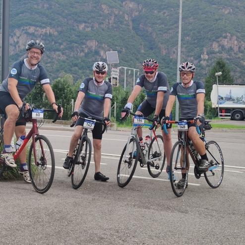 Fünf Radfahrer in Sportbekleidung stehen neben ihren Fahrrädern auf einer Straße vor einer bergigen Landschaft.