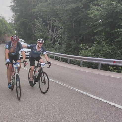 Zwei Radfahrer fahren auf einer kurvigen Straße, umgeben von Bäumen und einem steinigen Hang.