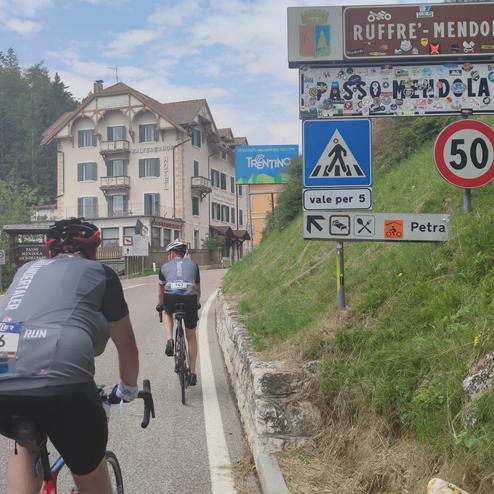 Zwei Radfahrer fahren auf einer Straße vorbei an einem Hotel und Verkehrsschildern zum Pass Mendola.