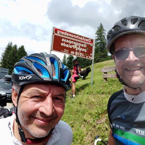Selbstporträt von zwei Radfahrern mit Helmen vor einem Schild, das auf den Deutenhauserpass hinweist. Verkehr im Hintergrund.