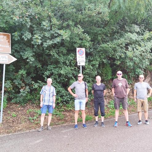 Fünf Personen stehen neben einem Wegweiser in der Natur, umgeben von Bäumen und einer Steinwand.
