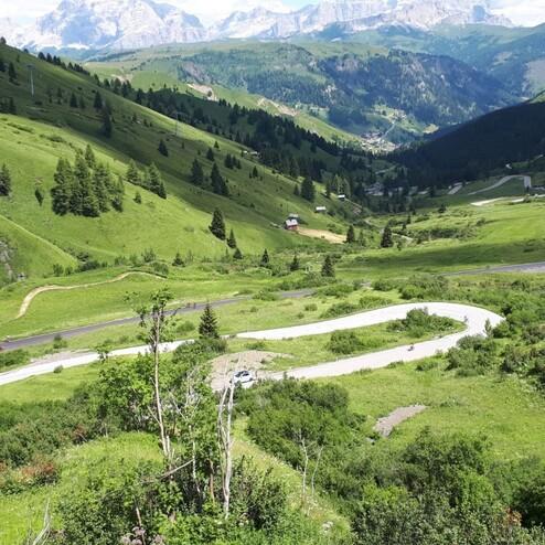 Panoramaaussicht auf eine grüne Berglandschaft mit kurvenreicher Straße und kleinen Hütten im Hintergrund.