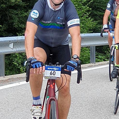 Radfahrer in Sportkleidung mit Helm und Sonnenbrille, fährt auf einer Straße, im Hintergrund weitere Radfahrer und Bäume.