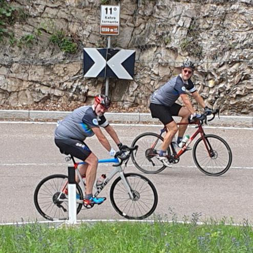 Zwei Radfahrer auf einer kurvigen Straße, umgeben von Felsen und einem Hinweis-Schild.