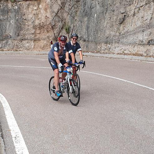 Zwei Radfahrer auf einer kurvigen Bergstraße, umgeben von felsiger Landschaft und blühendem Gras.