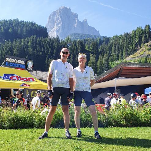 Zwei Radfahrer in Sportbekleidung posieren vor einer Bergkulisse und einer Ski-Schule in Sankt Cristina.