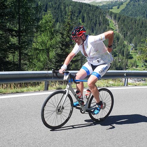 Radfahrer in Sportbekleidung fährt einen steilen Bergweg, mit Felsen und Bäumen im Hintergrund.