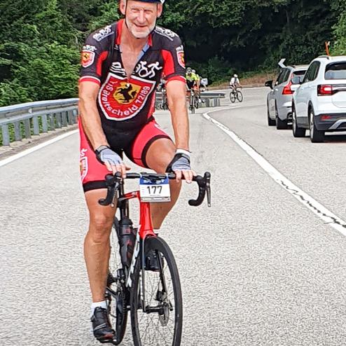Radfahrer in schwarzer und roter Sportkleidung fährt auf einer Landstraße, umgeben von grüner Vegetation.