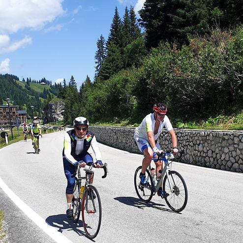 Zwei Radfahrer auf einer kurvigen Landstraße mit Bäumen und Bergen im Hintergrund, während andere Radfahrer vorbeifahren.