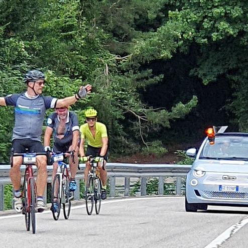 Drei Radfahrer auf einer Landstraße, einer davon mit ausgestreckten Armen, begleitet von einem Fahrzeug mit Warnleuchte.