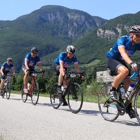 Sechs Radfahrer in blauen Trikots fahren auf einer Straße mit Bergen im Hintergrund.