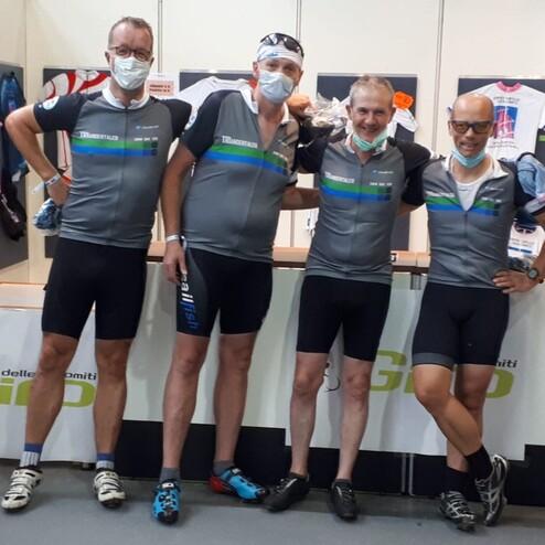 Vier Radfahrer in Sportbekleidung stehen lächelnd vor einem Verkaufsstand mit Trikots und Zubehör.