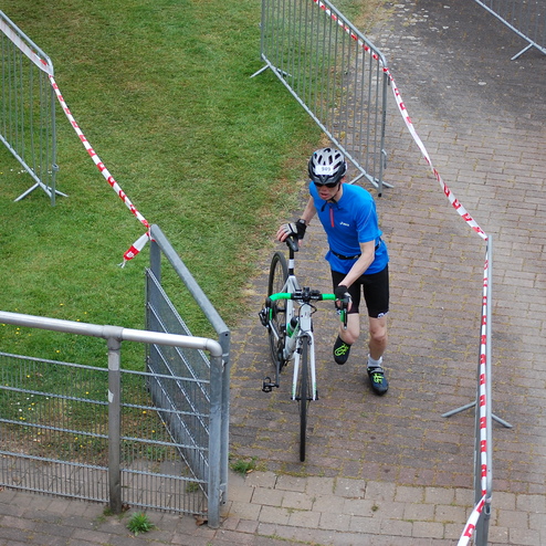 Radfahrer mit Helm zieht sein Fahrrad durch einen abgesperrten Bereich auf einer Wiese.