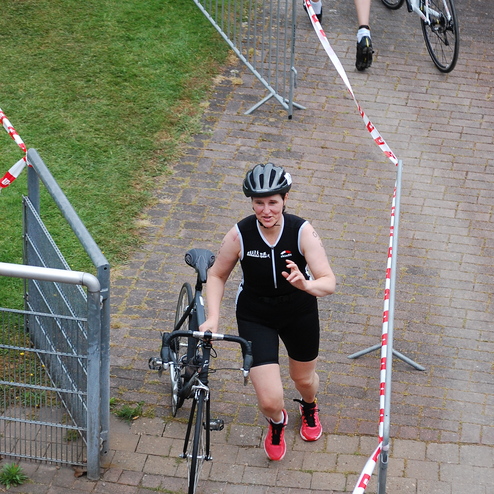 Frau in Sportkleidung trägt ein Fahrrad beim Triathlon-Wettbewerb auf einem gepflasterten Weg.