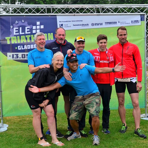 Gruppe von sieben Sportlern posiert vor einem Banner des ELE Triathlons in Gladbeck, umgeben von grüner Wiese.