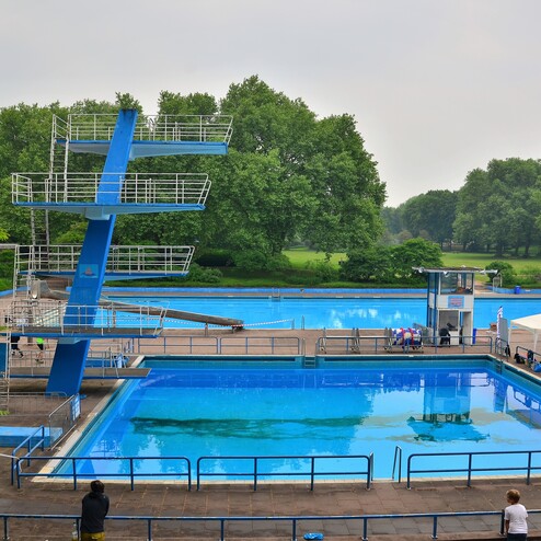 Blick auf ein Sprungbrett neben einem Schwimmbecken in einem Park, umgeben von Bäumen und einer Gruppe von Menschen.