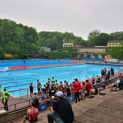 Sportveranstaltung am Schwimmbecken mit Teilnehmern, Zuschauern und einem historischen Gebäude im Hintergrund.