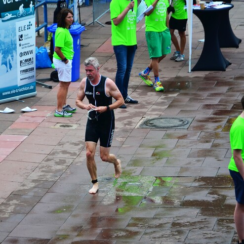 Mann in sportlicher Kleidung läuft barfuß auf nassem Boden, umgeben von Zuschauern in grünen T-Shirts.