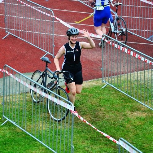 Triathletin mit Fahrrad, die winkt, in einem abgesperrten Bereich einer Sportveranstaltung auf einer roten Laufbahn.