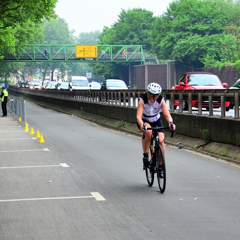 Radfahrerin in sportlicher Kleidung fährt auf einer Straße, umgeben von Bäumen und Verkehrssperren.