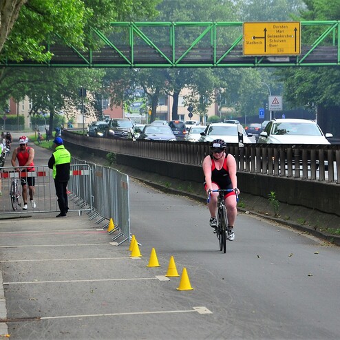 Zwei Radfahrer in Sportkleidung auf einem abgesperrten Weg neben einer stark befahrenen Straße. Verkehrshinweisschild im Hintergrund.