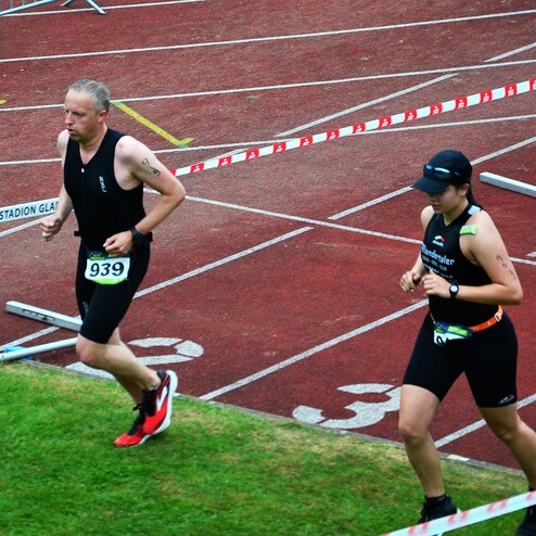 Zwei Athleten laufen auf einer roten Laufbahn, umrahmt von Absperrbändern und Hürden während eines Wettkampfs.