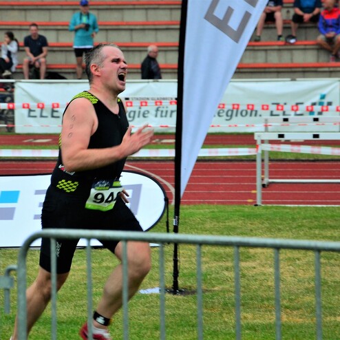 Ein Athlet in Wettkampfbekleidung sprintet auf einer Laufbahn, während Zuschauer auf den Tribünen zuschauen.