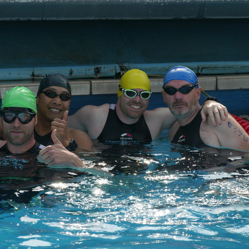 Vier Schwimmer in Badesachen posieren lächelnd im Wasser, während einer mit Daumen hoch die Kamera anzeigt.