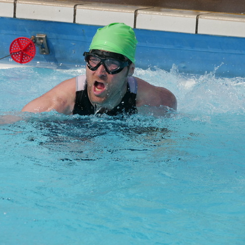 Schwimmer mit grüner Badekappe und Schwimmbrille kämpft im Wasser, Spritzer um ihn herum, rote Bojen im Hintergrund.