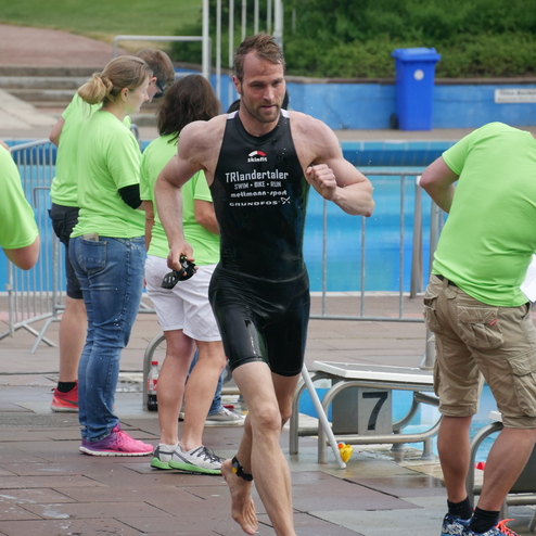 Triathlet in schwarzem Anzug sprintet nach dem Schwimmen aus dem Wasser, umgeben von Helfern in grünen T-Shirts.