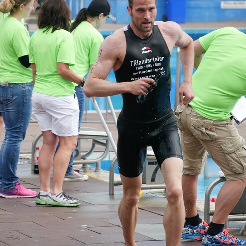 Ein Athlet in schwarzem Sportanzug läuft nass und barfuß entlang einer Schwimmbeckenrand mit zuschauenden Personen.