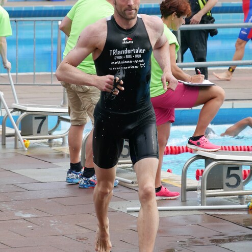 Ein Athlet in einem schwarzen Triathlonanzug läuft nass vom Schwimmbecken, umgeben von Zuschauern und Helfern.