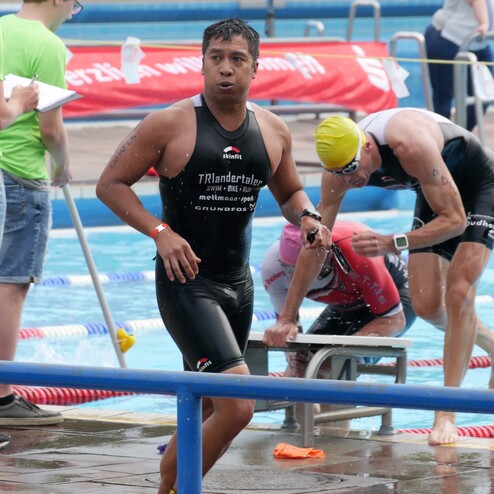 Triathlet in einem schwarzen Anzug verlässt das Wasser, während andere Athleten sich auf einen Schwimmstart vorbereiten.