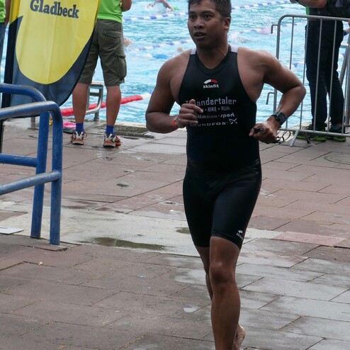 Ein Sportler in einem Neoprenanzug läuft barfuß entlang einer nassen Fläche nahe einem Schwimmbecken.