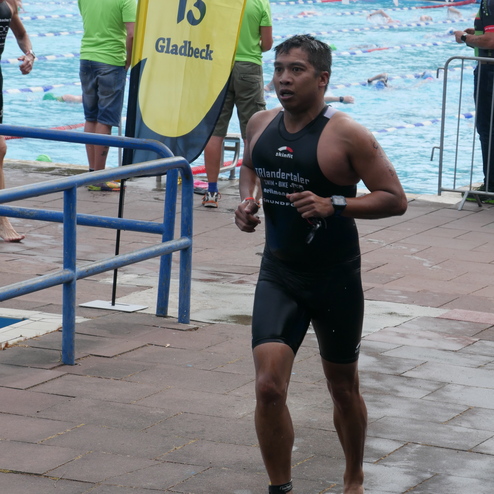 Triathlet in Neoprenanzug läuft barfuß an einem Schwimmbecken vorbei, umgeben von anderen Teilnehmern und Zuschauern.