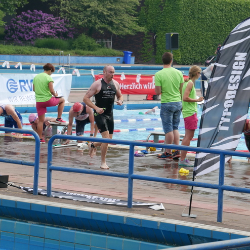 Triathlet verlässt das Schwimmbecken, während Zuschauer und Helfer am Beckenrand stehen. Hintergrund: Pflanzen und Banner.