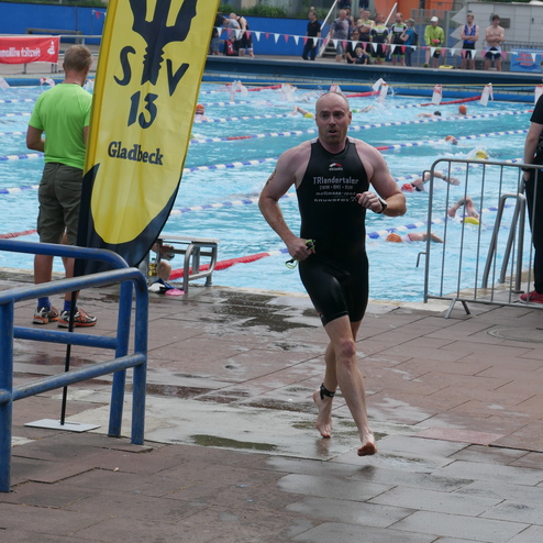 Ein Teilnehmer in Schwimmbekleidung läuft nach dem Schwimmen aus einem Poolbereich, umgeben von Zuschauern und Wettkämpfern.