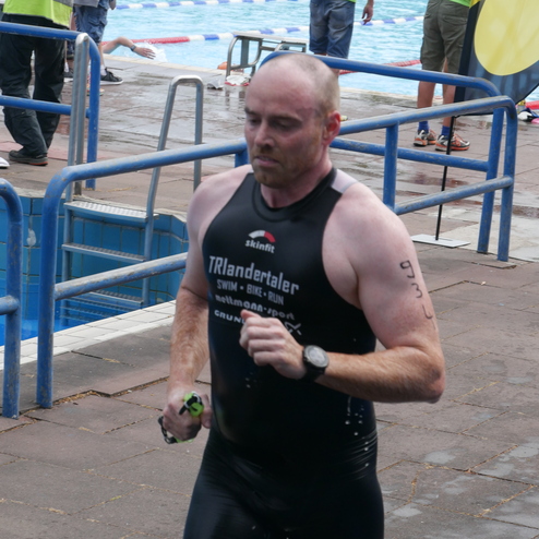 Ein Mann in einem schwarzen Triathlonanzug läuft am Beckenrand eines Schwimmbades während eines Wettkampfs.