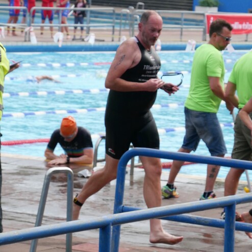 Ein Schwimmer in Neoprenanzug geht barfuß aus dem Wasser, im Hintergrund sind weitere Schwimmer und Trainer sichtbar.