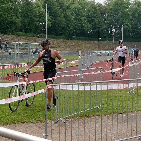 Teilnehmende Triathleten tragen ihre Fahrräder auf einem Wettkampfgelände, umgeben von Absperrungen und Zuschauern.