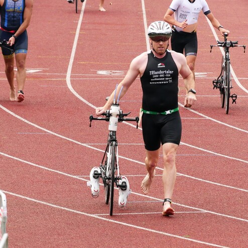 Triathlet trägt sein Fahrrad auf einer Tartanbahn, andere Athleten sind im Hintergrund sichtbar.