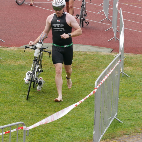 Triathlet läuft barfuß mit Fahrrad zur Wechselzone auf rutschigem Untergrund, andere Sportler im Hintergrund.