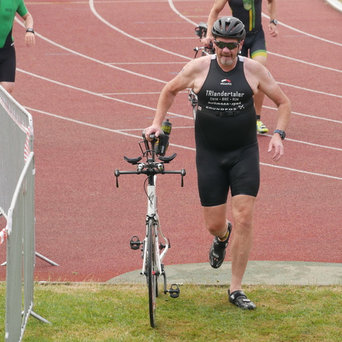 Mann in einem Triathlonanzug läuft mit Fahrrad über eine Rasenfläche neben einer Laufbahn. Weitere Teilnehmer im Hintergrund.