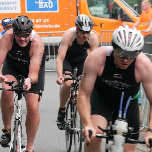 Drei Triathleten in Sportanzügen fahren rasant auf Fahrrädern während eines Wettkampfs auf einer Straße.