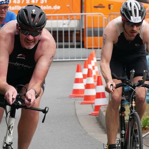 Zwei Triathleten auf Fahrrädern fahren an orange-weißen Kunststoffconen vorbei, einer trägt eine Startnummer auf dem Helm.