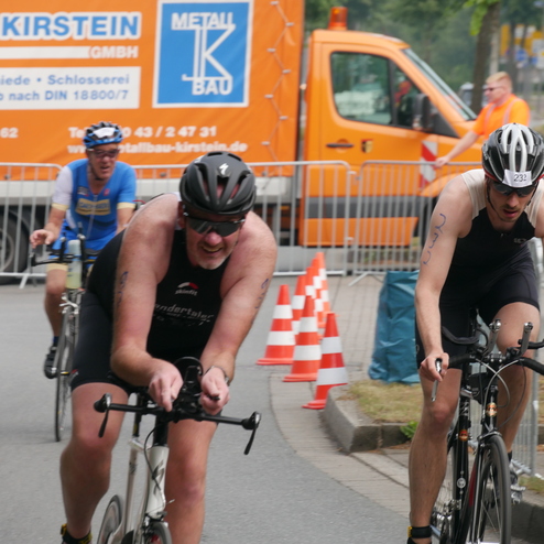 Zwei Radfahrer fahren in einem Wettkampf, im Hintergrund eine orangefarbene Nutzfahrzeug und Sicherheitsbarrieren.