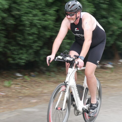Mann in Sportbekleidung fährt auf einem Rennrad auf einer Straße, umgeben von grünen Büschen im Hintergrund.