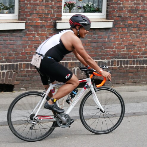 Radfahrer in Wettkampfkleidung fährt auf einem Straßenrad entlang einer Ziegelsteinmauer. Nummerntuch am Oberkörper sichtbar.
