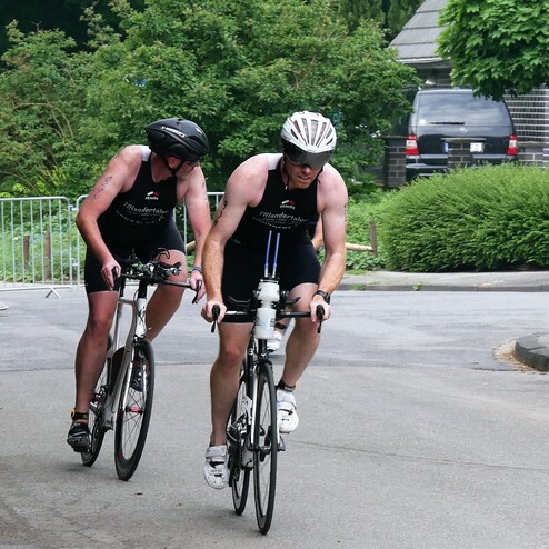 Zwei Männer in Triathlonanzügen fahren auf Rennrädern eine Straße entlang, ein Zuschauer sitzt im Hintergrund.