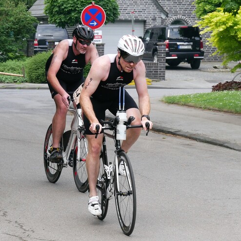 Zwei Athleten in Sportkleidung fahren auf Rennrädern eine Straße entlang, umgeben von grüner Natur und Wohnhäusern.